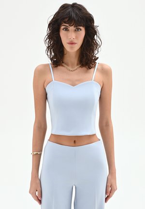 Donna con capelli ricci scuri che indossa un top corto celeste, pantaloni a vita alta abbinati, collana d'oro e braccialetto d'oro, in piedi dritta.