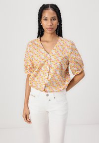 Blusa floreale a maniche corte in arancione, rosa e bianco. Collo a V con bottoni lungo la parte frontale. Indossata con jeans bianchi, che presentano una vestibilità rilassata.