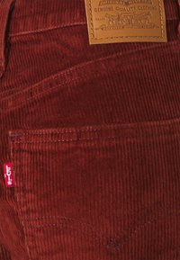 Pantalon en velours côtelé couleur rouille avec une pièce en cuir portant le logo de la marque, des coutures apparentes et un design classique des poches.