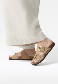 Sandales mules en daim marron avec deux boucles réglables, semelle en liège, semelle en caoutchouc texturée et design à bout ouvert, assorties à un pantalon en tissu léger.
