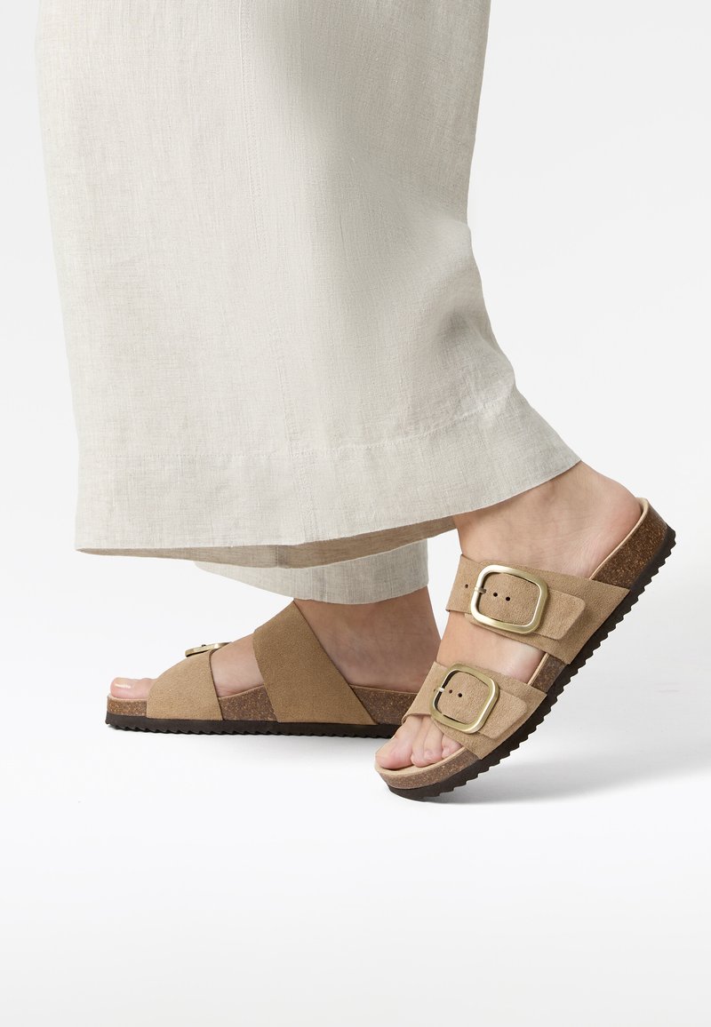Sandales mules en daim marron avec deux boucles réglables, semelle en liège, semelle en caoutchouc texturée et design à bout ouvert, assorties à un pantalon en tissu léger.