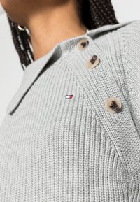 Maglione grigio lavorato a maglia con design a colletto, grandi bottoni beige e un piccolo logo con accenti rossi e blu sul tessuto.