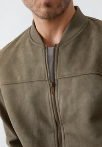 Mężczyzna w oliwkowej skórzanej kurtce z metalowym zamkiem, częściowo rozpiętej, ukazującej białą koszulę pod spodem, przyciętej na wysokości brody i klatki piersiowej.