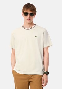 T-shirt beige clair avec un col rond côtelé orné de rayures rouges et marron, manches courtes, et un logo crocodile vert sur la poitrine.
