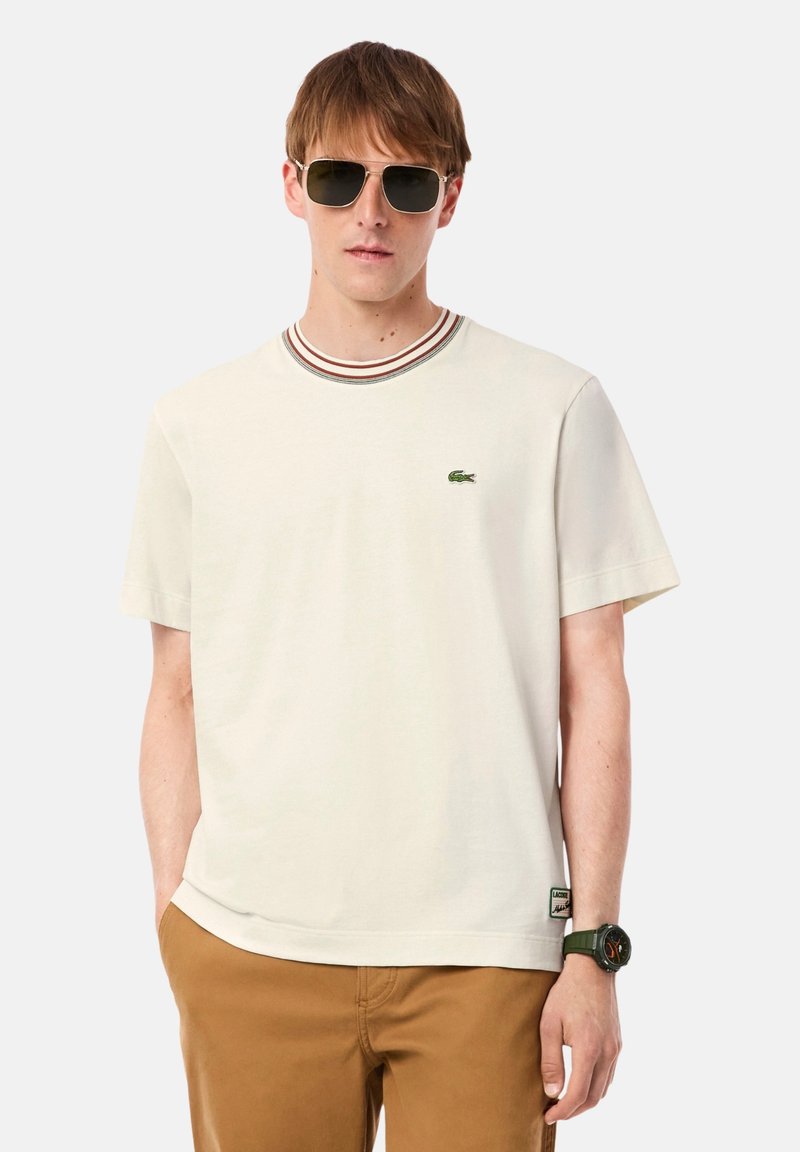 T-shirt beige clair avec un col rond côtelé orné de rayures rouges et marron, manches courtes, et un logo crocodile vert sur la poitrine.