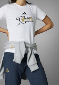 Valkoinen t-paita, jossa musta ja kultainen teksti 50. BMW Berlin Marathonista, yhdistettynä laivastonsinisiin housuihin ja harmaaseen collegepaitaan, joka on sidottu vyötärölle.