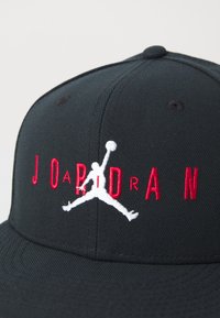 Sort kasket med rød "JORDAN" tekst og hvid silhuet af en basketballspiller, der hopper med bolden i midten af luften på frontpanelet.