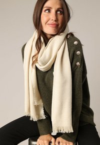 Écharpe couleur crème avec un tissage doux et texturé, drapée autour du cou, complétant un pull vert olive avec des boutons décoratifs.