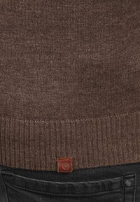 Brun strikket sweater med ribbet kant og et lædermærke forneden. Teksturen er glat og ensartet, hvilket komplementerer mørke jeans.