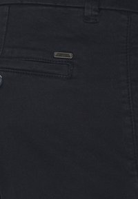 Dunkelblaue Jeans aus Denim verfügen über eine seitliche Tasche und ein kleines schwarzes Markenlabel. Der Stoff hat eine glatte, leicht strukturierte Oberfläche.