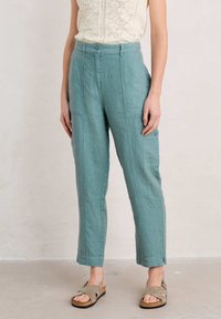 Pantalons en lin bleu clair avec une coupe droite, poches avant et détail de bouton sur le côté. Assortis à des sandales ouvertes grises.