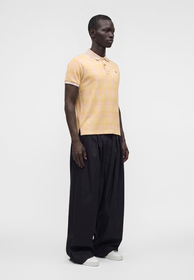 Homme portant un polo jaune pâle à manches courtes, un pantalon large noir et des baskets blanches, debout devant un fond clair uni.