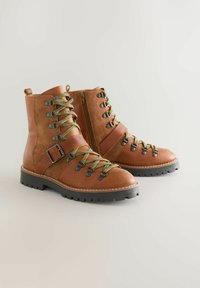 Braune Wanderstiefel aus Leder mit strukturiertem Obermaterial, metallischen Details, stofflichen Schnürsenkeln in Gelb und Grün sowie robusten schwarzen Sohlen. Seitlicher Reißverschluss.