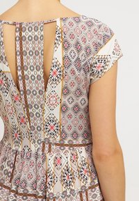 Robe en tissu à motif beige, ornée de dessins géométriques en rouge, noir et or. Le dos présente un décolleté en V profond avec un détail en forme de trou clé.