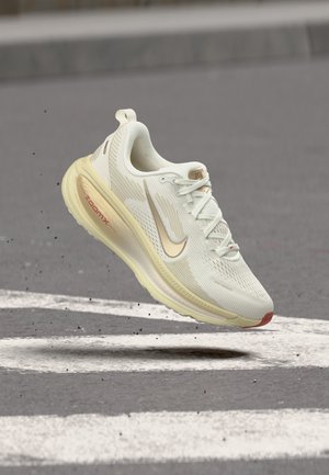 Hellbeiger Nike ZoomX Laufschuh, der über einer grauen Asphaltstraße mit weißen Markierungen schwebt.