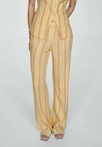 Persoon in gele en witte verticaal gestreepte brede broek en bijpassende mouwloze vest, met beige open teen sandalen met vierkante hak.