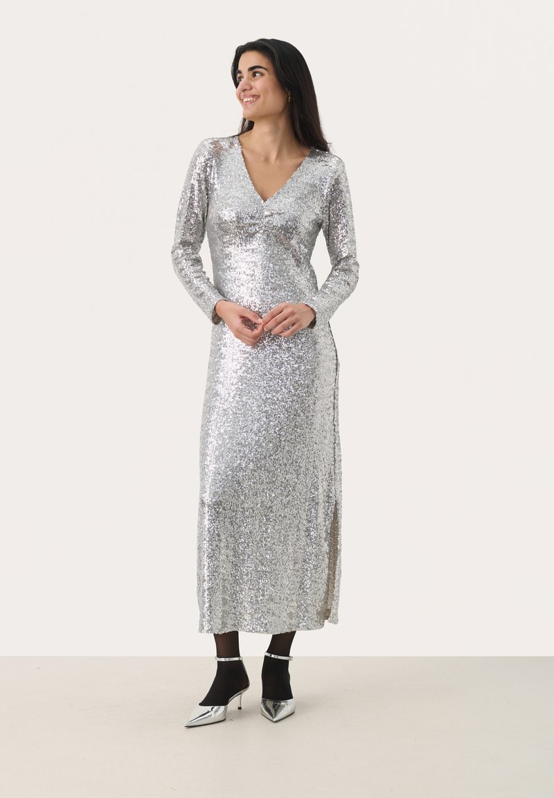 Silbernes Paillettenkleid mit langen Ärmeln und V-Ausschnitt. Es hat ein tailliertes Oberteil und einen Seitenschlitz, ergänzt durch metallische High Heels mit spitzem Zehenbereich.