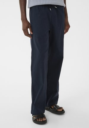 Personne portant un pantalon ample bleu marine avec une taille à cordon et des sandales noires à bout ouvert, debout sur un fond blanc uni.