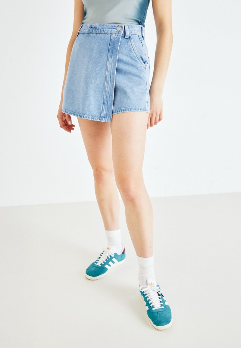 Persona vestida con una falda envolvente de mezclilla azul claro, zapatillas de deporte turquesa con rayas blancas, calcetines blancos y una camiseta sin mangas verde, de pie sobre un suelo blanco.