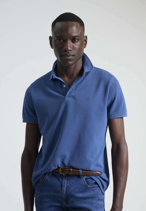 Camisa polo azul con cuello y mangas cortas. Presenta una tela texturizada y un pequeño logo en el pecho. Se lleva con jeans de mezclilla.