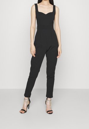 Schwarzer Jumpsuit mit quadratischem Ausschnitt, ärmellosem Design und schmal zulaufenden Beinen. Kombiniert mit schwarzen High-Heelsandalen mit Perlenverzierung.