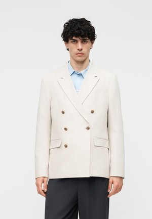 Ung mann iført en kremfarget dobbelspent blazer med brune knapper, lyseblå skjorte og mørke bukser, stående foran en hvit bakgrunn.