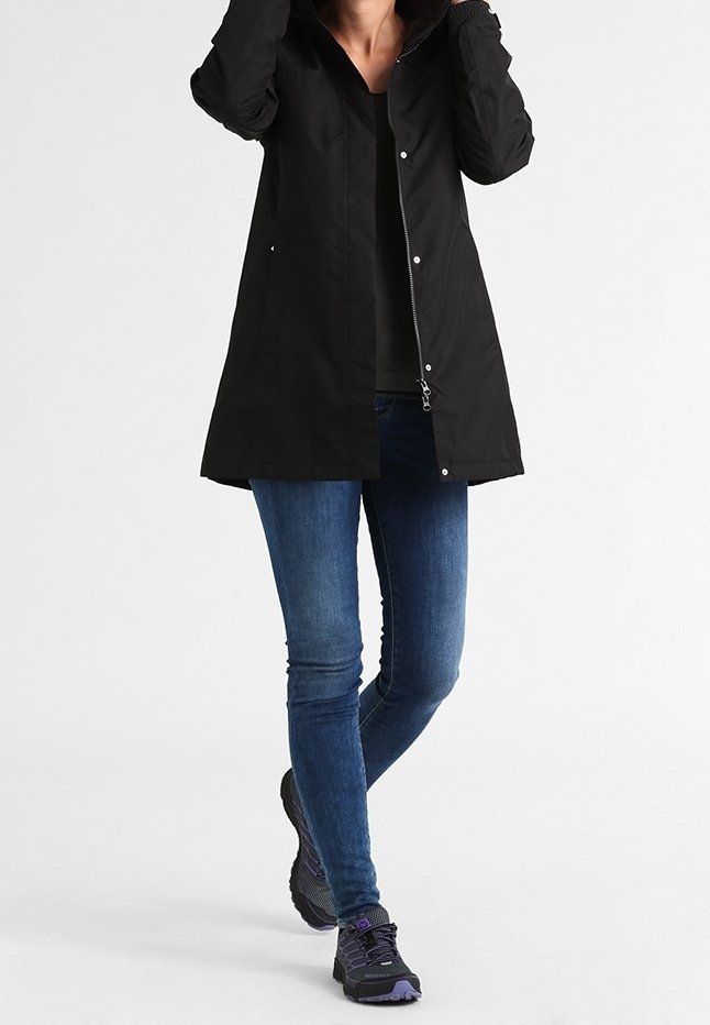 Personne portant une veste noire, un jean skinny bleu et des baskets noires marchant avec les mains près du col de la veste sur un fond uni.