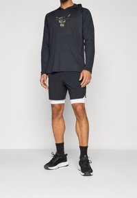 Schwarzer Kapuzenpullover mit langen Ärmeln und einem Camouflage-Bullenlogo, kombiniert mit schwarzen Shorts mit weißem Saum und schwarzen Sportschuhen vor einem neutralen Hintergrund.