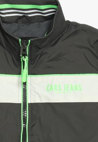 Chaqueta de cremallera en blanco y negro con acentos en verde neón, cuello acanalado a rayas y logo "CARS JEANS" en el pecho.