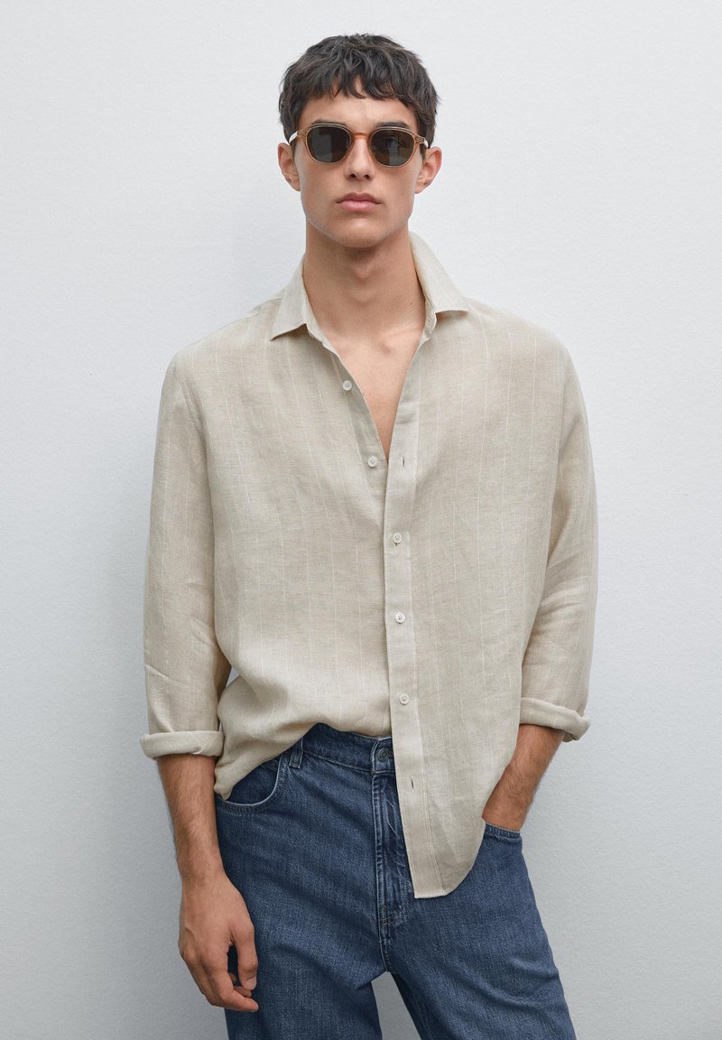 Young man wearing round brown sunglasses, beige button-up shirt with rolled sleeves, and blue jeans, standing against plain light wall.