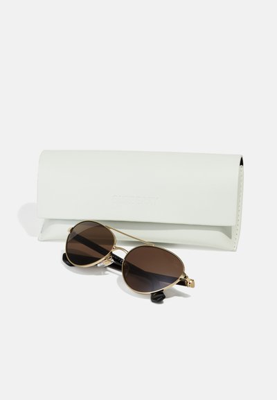 Sonnenbrille mit goldenem Metallrahmen und dunklen Gläsern, liegend vor einem weißen rechteckigen Brillenetui auf weißem Hintergrund.
