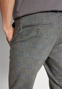 Pantalons à carreaux gris en tissu texturé, comportant une poche arrière boutonnée et une subtile touche de bleu dans le motif.