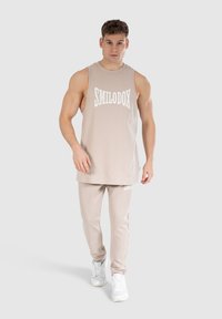 Beige tanktop med hvid "SMILODOX" logo, parret med matchende beige joggers og hvide sneakers. Glat stof, afslappet pasform.