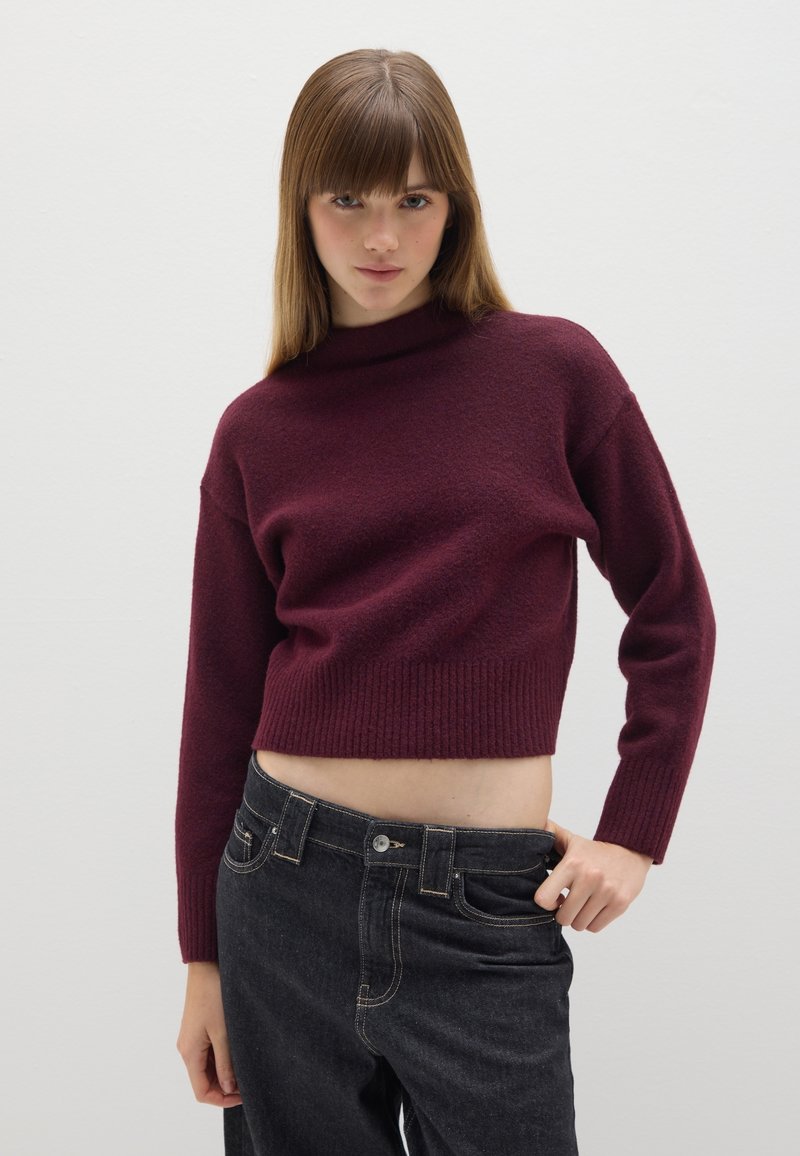 Jonge vrouw met steil bruin haar, gekleed in een korte bordeauxrode trui en zwarte hoge taille jeans, tegen een effen witte achtergrond.