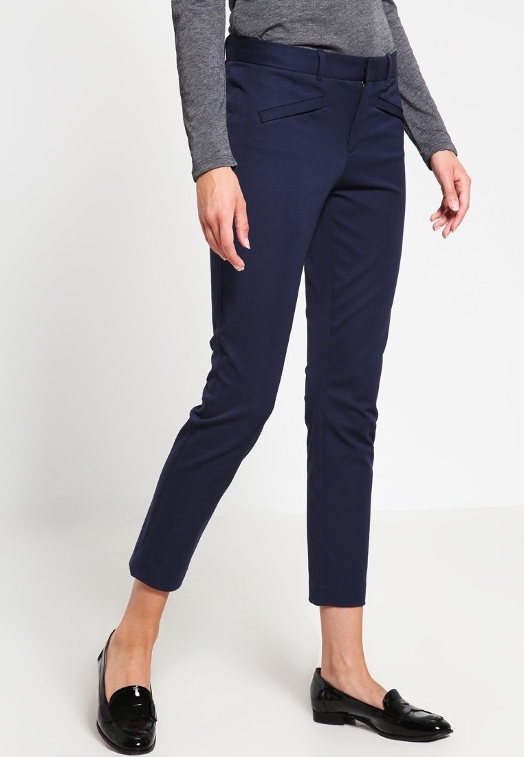 Femme portant un pantalon slim bleu marine raccourci, un haut gris à manches longues et des mocassins noirs brillants, debout devant un fond blanc.