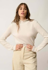 Weiße, gerippte Rollkragensweater mit einem Rüschensaum. Weicher Stoff und enganliegendes Design, kombiniert mit hellbeigen Cordhosen mit vertikalen Streifen.