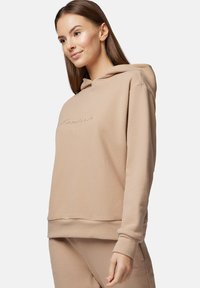 Beżowa bluza z kapturem o luźnym kroju, miękkiej fakturze i wyszywanym logo z przodu. Posiada kieszeń kangurka i ściągacze na mankietach.
