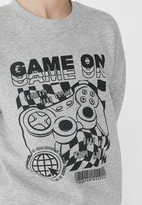 Šedá mikina s velkým černým nápisem "HRA ZAČÍNÁ", grafikou herního ovladače a kostkovanými akcenty. Jednoduchý, pohodlný design.