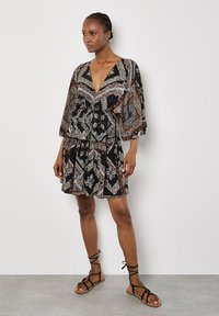 Robe noire avec un décolleté en V profond, des manches fluides et des motifs paisleyintriqués en blanc et marron. Associée à des sandales noires à lacets.