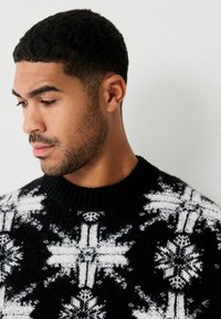 Pull en tricot noir avec un motif de flocon de neige blanc, col rond et poignets côtelés. Le tissu texturé apporte de la profondeur au design.