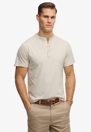 Man met beige henley-shirt met korte mouwen, bruine leren riem en beige broek met handen in de zakken tegen een witte achtergrond.
