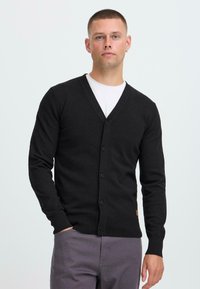 Man med kort hår som bär en svart knappad kavaj över en vit skjorta, står med en hand i fickan mot en neutral bakgrund.