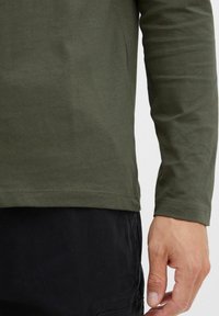 Olive green long-sleeve shirt in smooth cotton fabric, featuring a rounded hem and contrasting black pants, with visible stitching details.