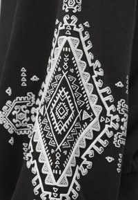 Melns audums ar sarežģītu baltu ģeometrisko izšuvumu, kurā ir dimanta formas un stūrveida raksti, radot teksturētu vizuālo detaļu.