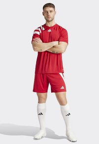 Červený športový dres a krátke nohavice s bielymi akcentmi a tromi pruhmi. Model nosí biele ponožky a kopačky, má skrížené ruky a stojí pred jednofarebným pozadím.