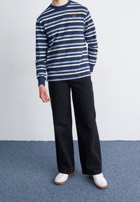 Musta raidallinen pitkähihaine paita, jossa on vaaleansinisiä ja mustia raitoja, varustettuna pienellä logolla. Yhdistetty mustiin leveisiin housuihin ja valkoisiin lenkkareihin.