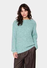 Šviesiai mėlynos spalvos megztinis su tekstūruotu raštu, apvaliu iškirpimu ir laisvomis rankovėmis, derinamas su rudos spalvos raukinėtomis kelnėmis.