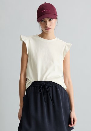 Kobieta ubrana w biały, bezrękawnik z marszczonymi ramionami, czarną spódnicę ze ściągaczem i bordową czapkę z różowym haftowanym napisem.