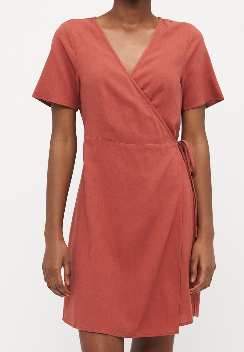 Femme portant une robe portefeuille manches courtes de couleur rouille avec un col en V et un détail de nœud sur le côté, sur fond blanc uni.