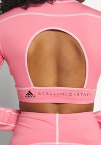 Rosa åtsittande träningsöverdel med utskärning på baksidan, med kontrasterande sömmar och svart varumärkestext, tillverkad av stretchigt och mjukt material.